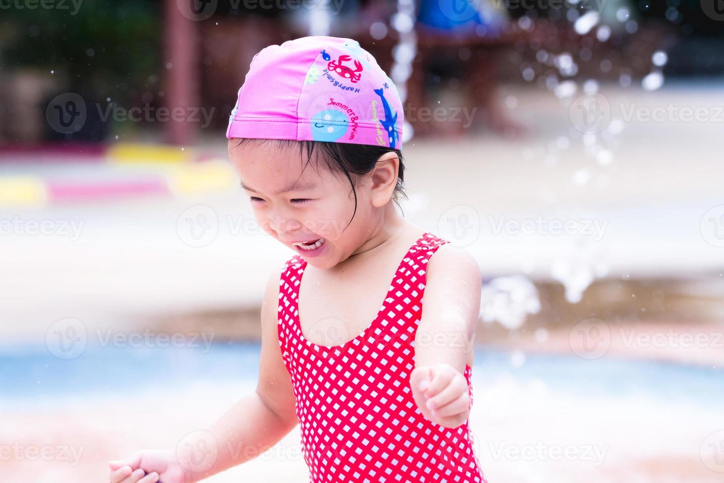 fille d'enfant asiatique heureux jouant de l'eau. rire des enfants. les enfants courent dans la fontaine. bébé portait un bonnet de bain rose et un maillot de bain rouge à losanges. bambin de 3 ans. photo