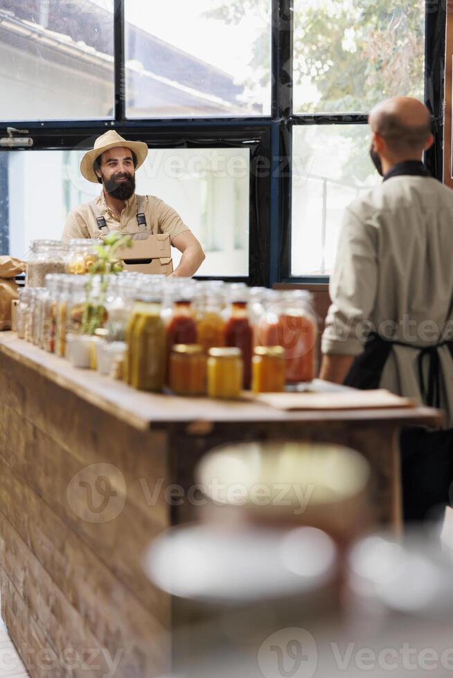 souriant homme portant une chapeau, entre dans éco amical épicerie boutique tandis que porter des boites de biologique des produits. Masculin vendeur avec noir tablier attendre à recevoir Frais paquets de caucasien agriculteur. photo