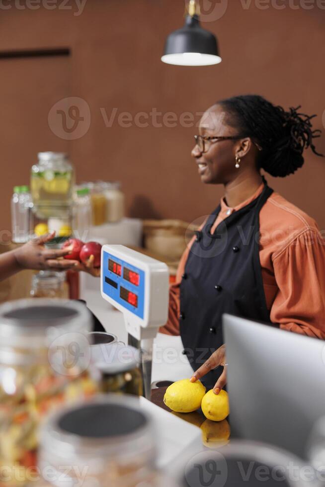 noir femme assiste une client dans une épicerie magasin, promouvoir localement grandi et biologique des produits. elles ou ils communiquer et utilisation La technologie pour un efficace check-out processus dans le respectueux de la nature marché. photo