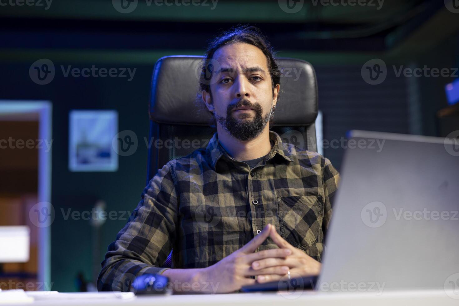 portrait de une gentilhomme assise à une bureau avec le sien mains franchi et à la recherche à caméra. sérieux Masculin individuel travail de maison, vérification e-mails, recherche sur le sien portable. photo