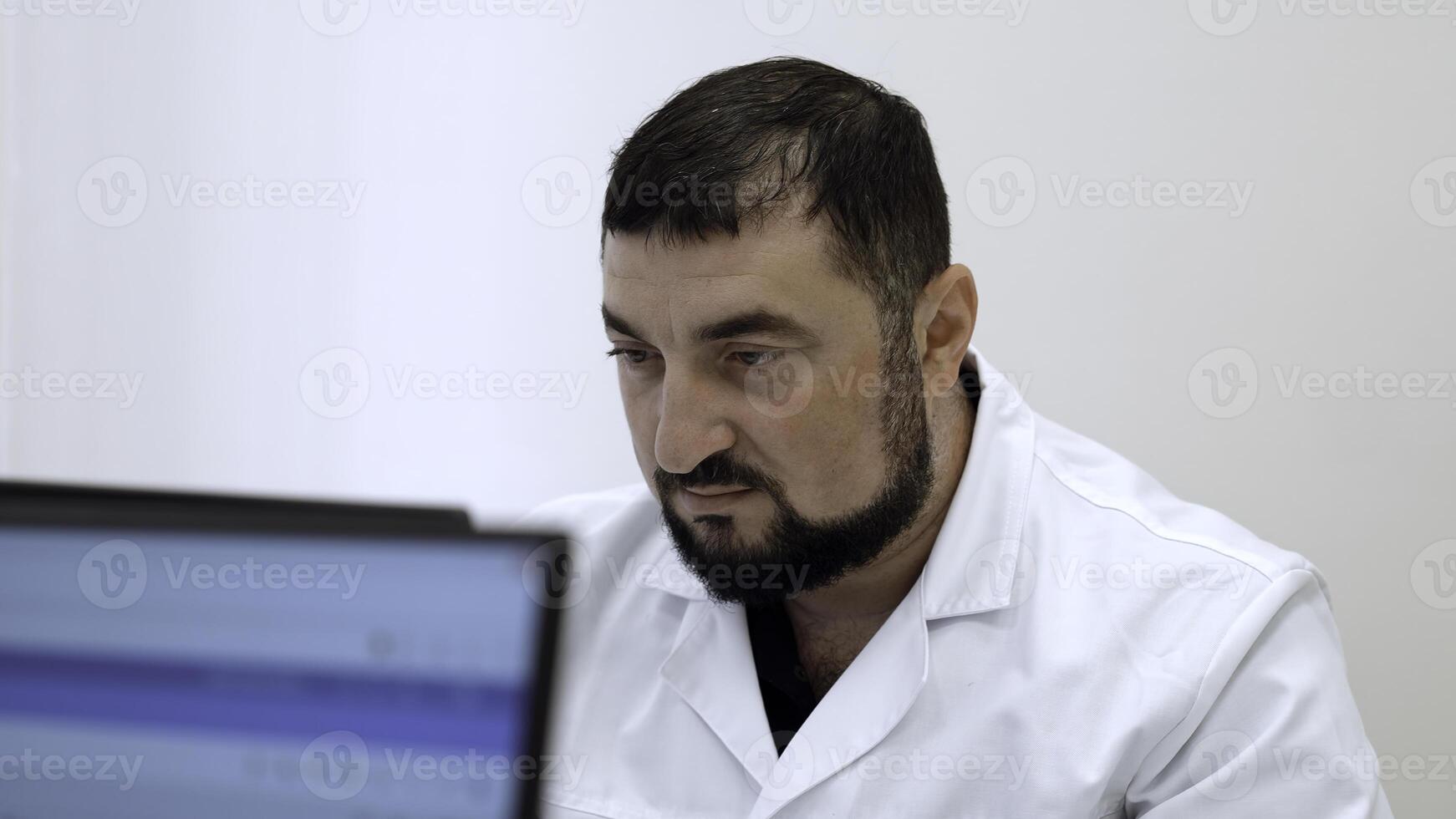 milieu vieilli médecin avec dans clinique. agrafe. barbu homme dans blanc manteau à la recherche à ordinateur moniteur. photo