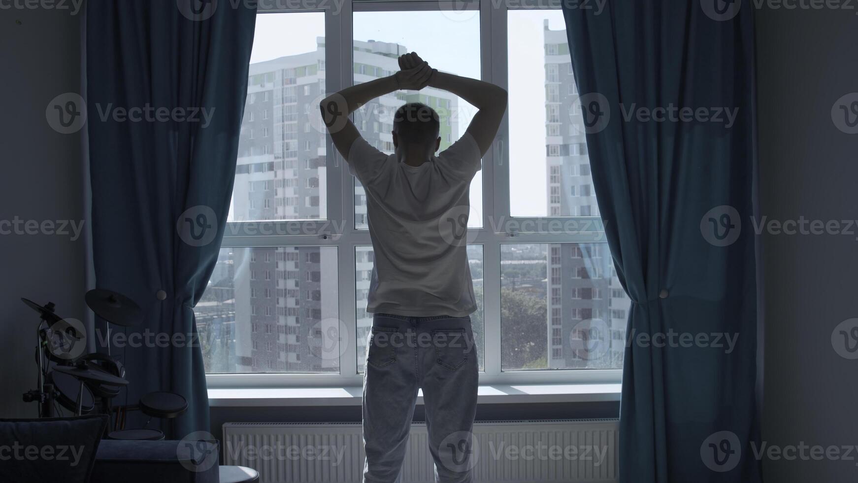 homme dans le Matin élevage le sien mains et élongation. médias. Jeune homme à la recherche par le fenêtre sur ville rue et bâtiments. photo