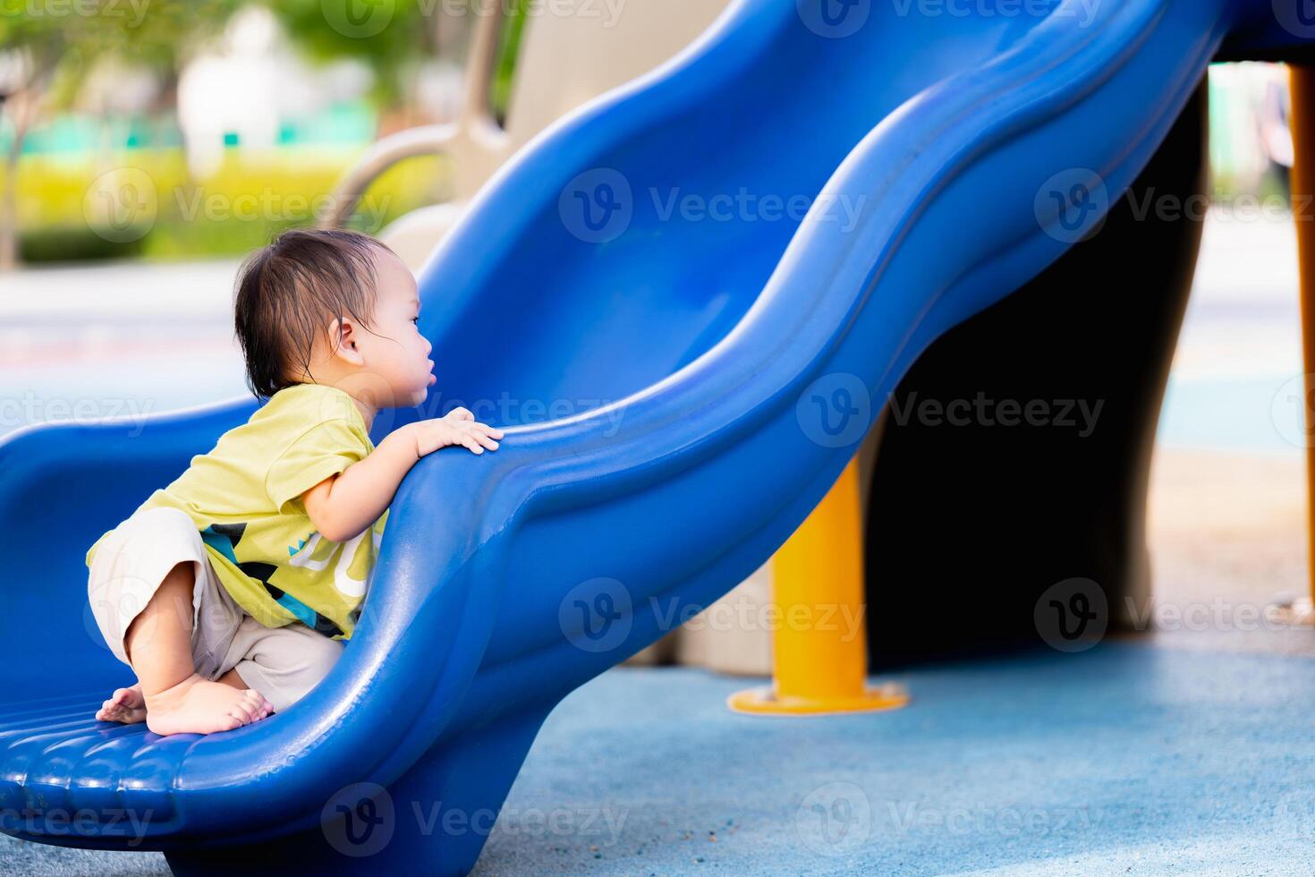 portrait de un asiatique bébé garçon en jouant sur une terrain de jeux glisser, fils séance à le fin de une bleu glisser, enfant apprentissage avec amusant, dans été ou printemps, un année vieux bambin avec apprendre et développer muscles. photo