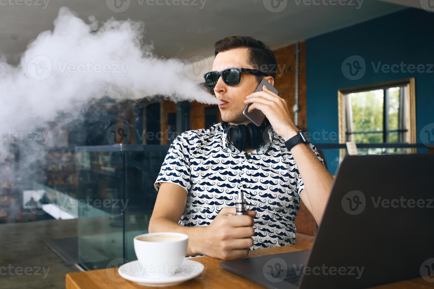 Jeune Beau branché homme lunettes de soleil séance dans café avec une tasse de café, vapoter et communiqués une nuage de vapeur. travail à ordinateur portable, parlant mobile téléphone et ayant une peu Pause. avec copie espace. photo