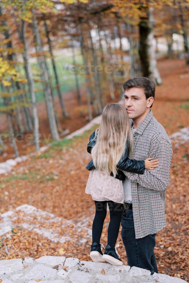 papa câlins une peu fille permanent sur une pierre chemin sur une colline dans le l'automne forêt et regards dans le distance photo