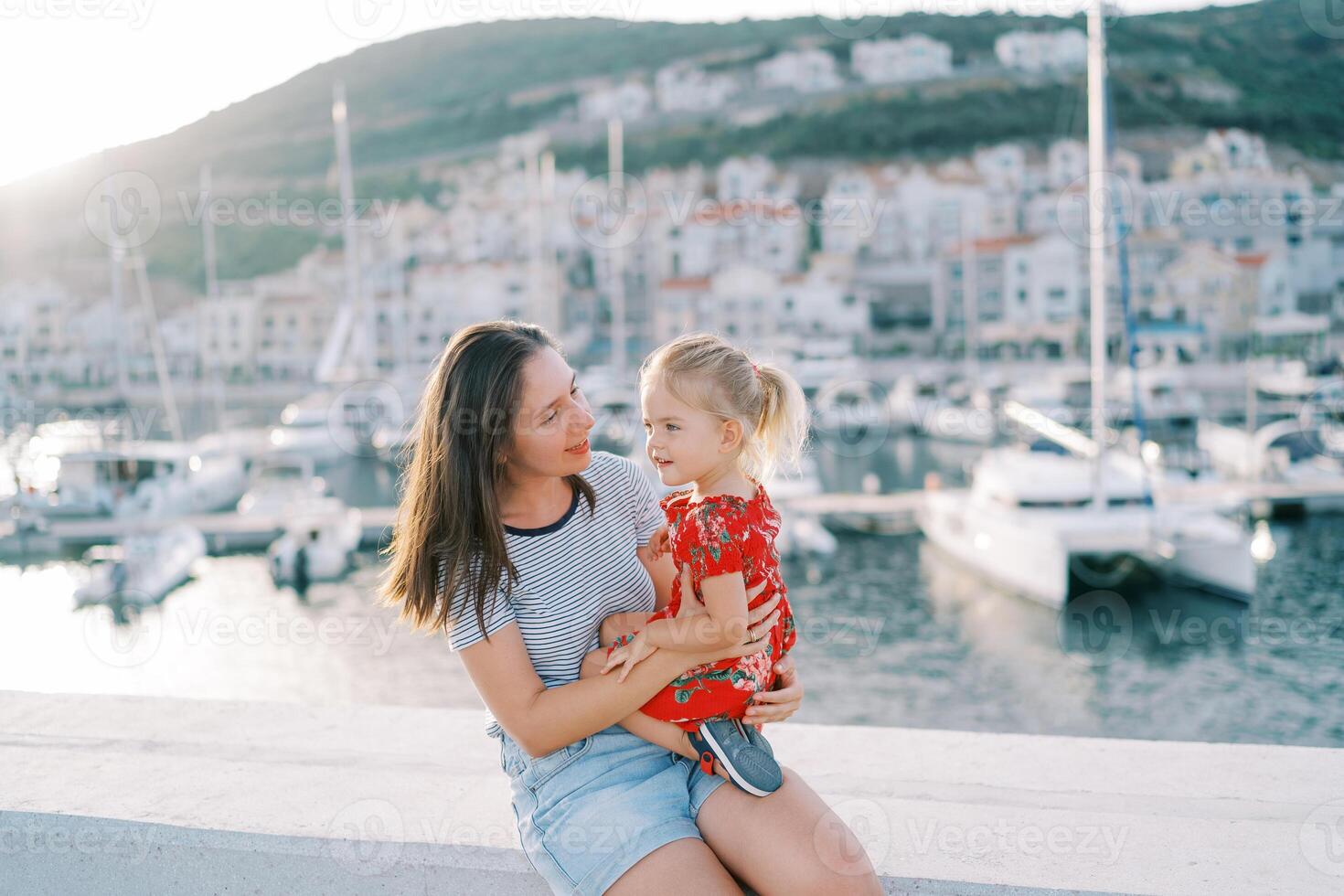 maman avec une peu fille dans sa bras est assis sur le clôture de le digue avec amarré yachts photo