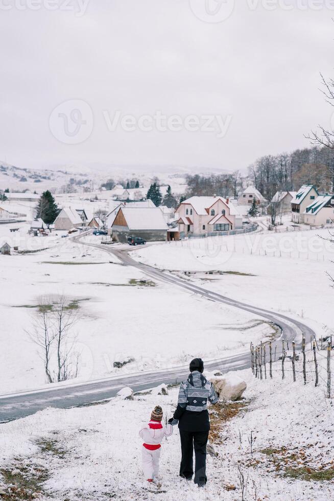 mère et une peu fille marcher vers le bas une neigeux colline en portant mains vers une village route. retour vue photo