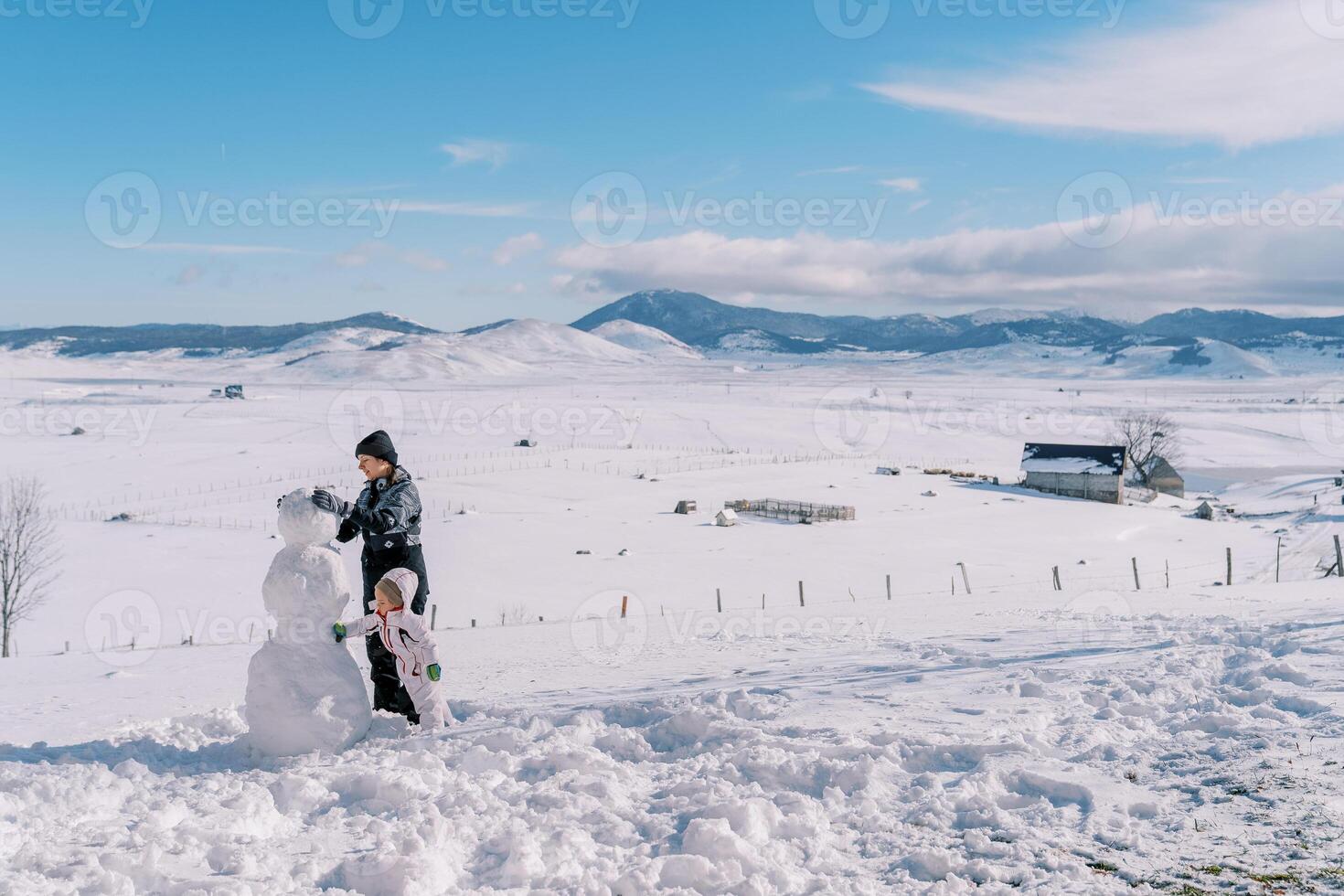 peu fille aide sa mère construire une bonhomme de neige dans une Montagne vallée. côté vue photo