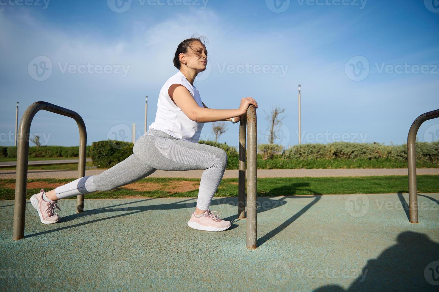 déterminé des sports femme dans blanc T-shirt et gris leggings, chauffage en haut, Faire fentes pour élongation jambe muscles. photo