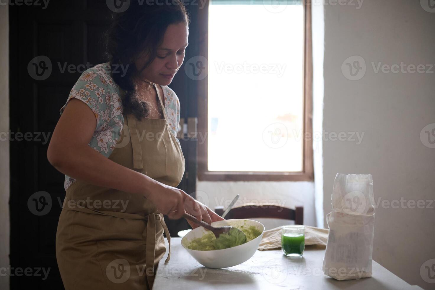 authentique femelle chef cuisinier dans beige tablier, en utilisant en bois cuillère, mélange épinard l'eau avec entier grain farine pour fabrication pâte, permanent à marbre cuisine table avec ingrédients, dans le campagne maison photo