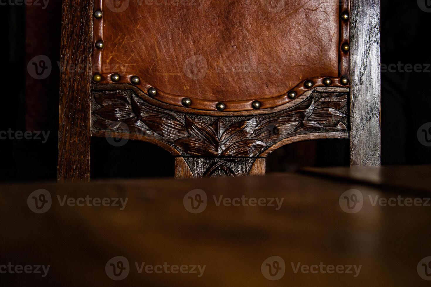 proche en haut de le détail de antique en bois chaise couvert avec marron cuir tapisserie, décoré avec métal rivets et artistique bois sculpture éléments. foncé Contexte. copie espace. photo