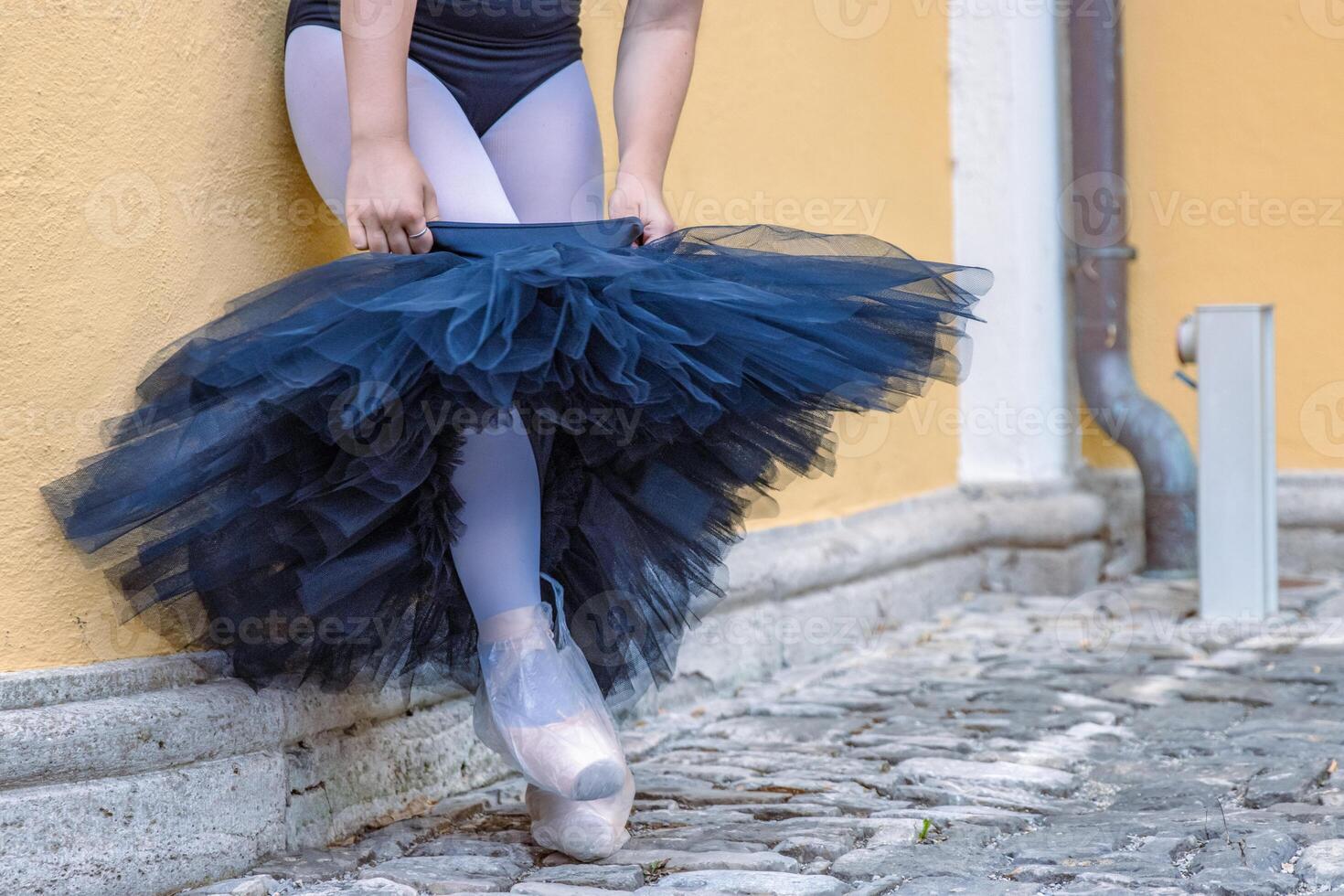 proche en haut Jeune femelle ballerine pansement en haut et en mettant sur noir tutu penché sur le Jaune bâtiment mur. ballet Danseur prise de le jupe. pointe des chaussures avec Plastique Sacs sur. noir cygne. photo