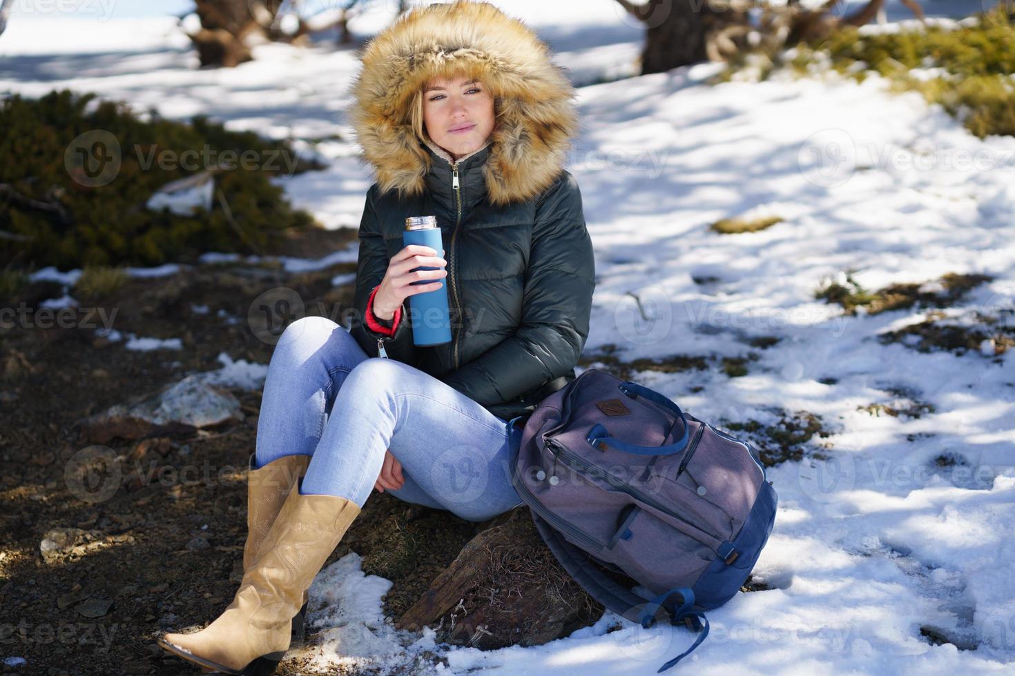 femme buvant quelque chose de chaud dans une bouteille thermos en métal assise sur un rocher dans les montagnes enneigées. photo