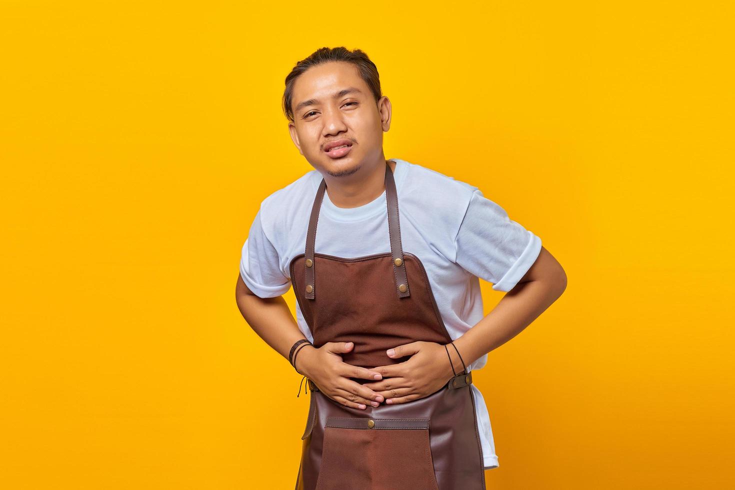 portrait d'un jeune homme asiatique portant un tablier montrant les mains sur l'estomac en raison de nausées, d'une maladie douloureuse se sentant mal photo