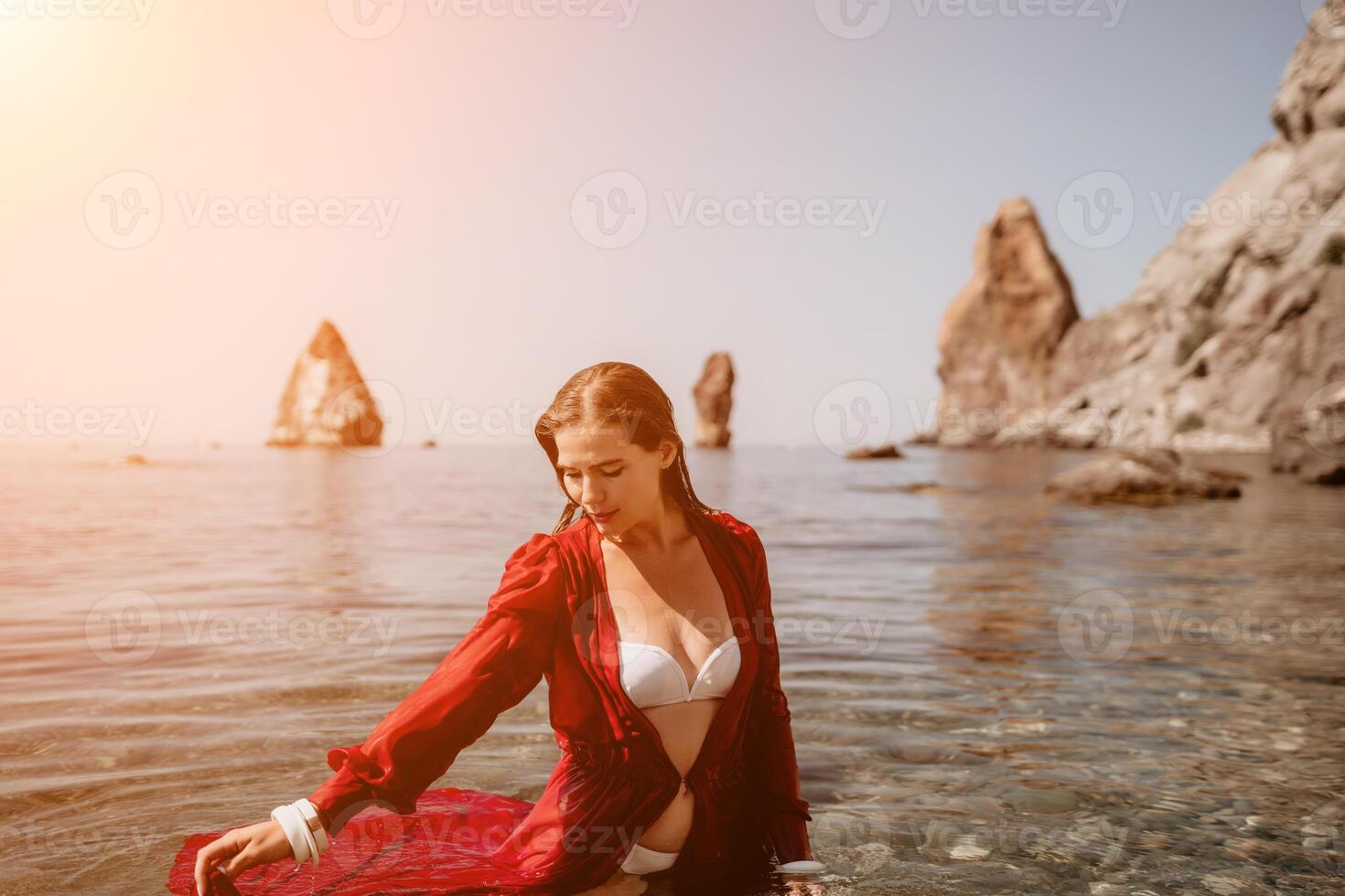 femme Voyage mer. content touristique dans rouge robe prendre plaisir prise image en plein air pour souvenirs. femme voyageur posant dans mer plage, entouré par volcanique montagnes, partage Voyage aventure périple photo