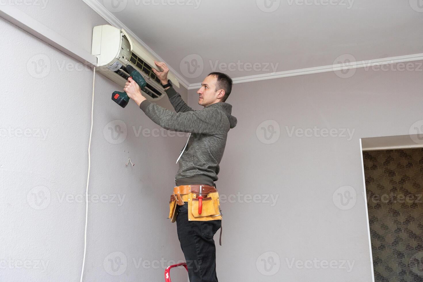 professionnel électricien homme avec Tournevis maintenir, nettoyage moderne air Conditionneur à l'intérieur. Jeune technicien permanent sur une échelle dans le pièce réparer ou installation air Conditionneur. photo