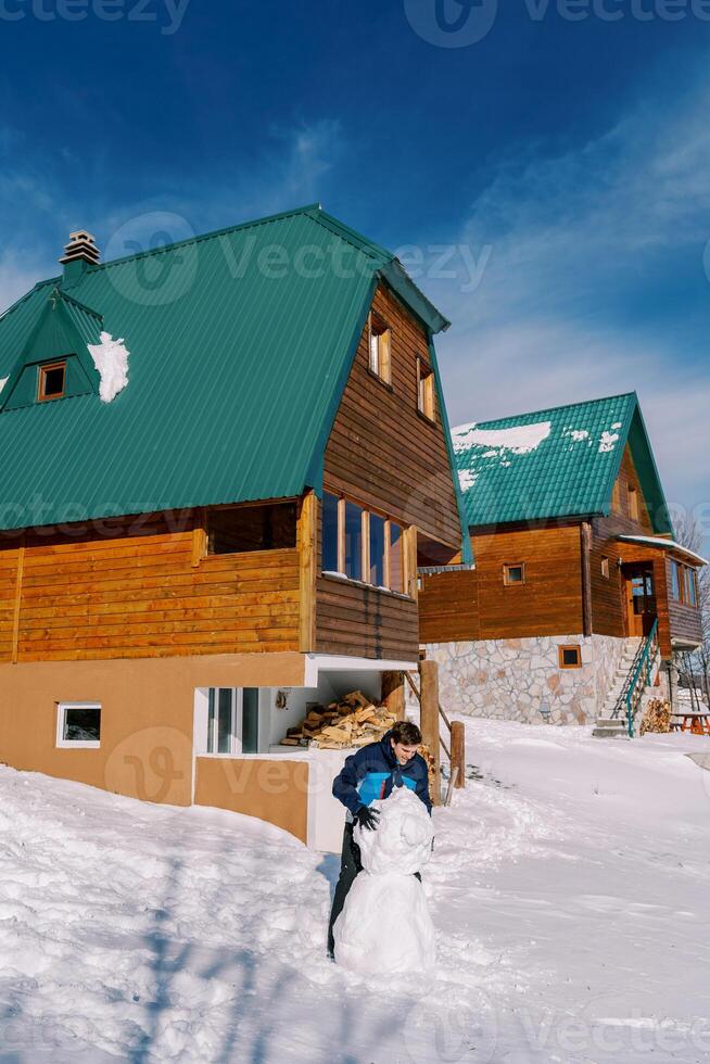 Jeune homme fait du une bonhomme de neige dans le Cour suivant à le en bois chalets photo