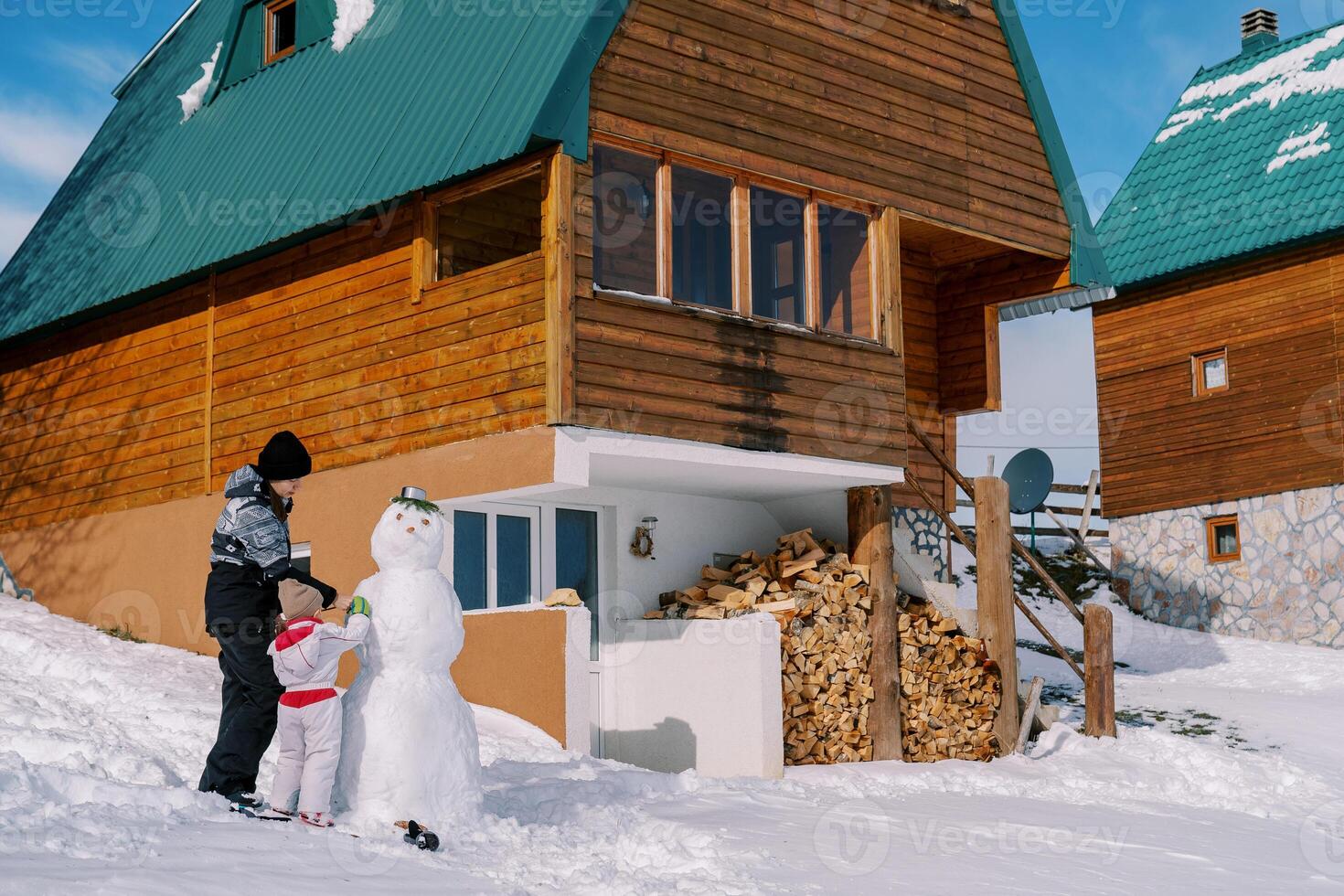 maman et peu fille décorer une bonhomme de neige près une tas de bois de une en bois chalet dans le neige photo