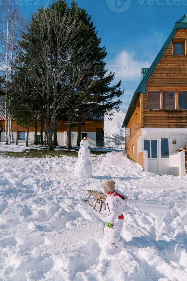 petit enfant tire une traîneau sur une corde passé une en bois chalet, à la recherche retour à le bonhomme de neige photo