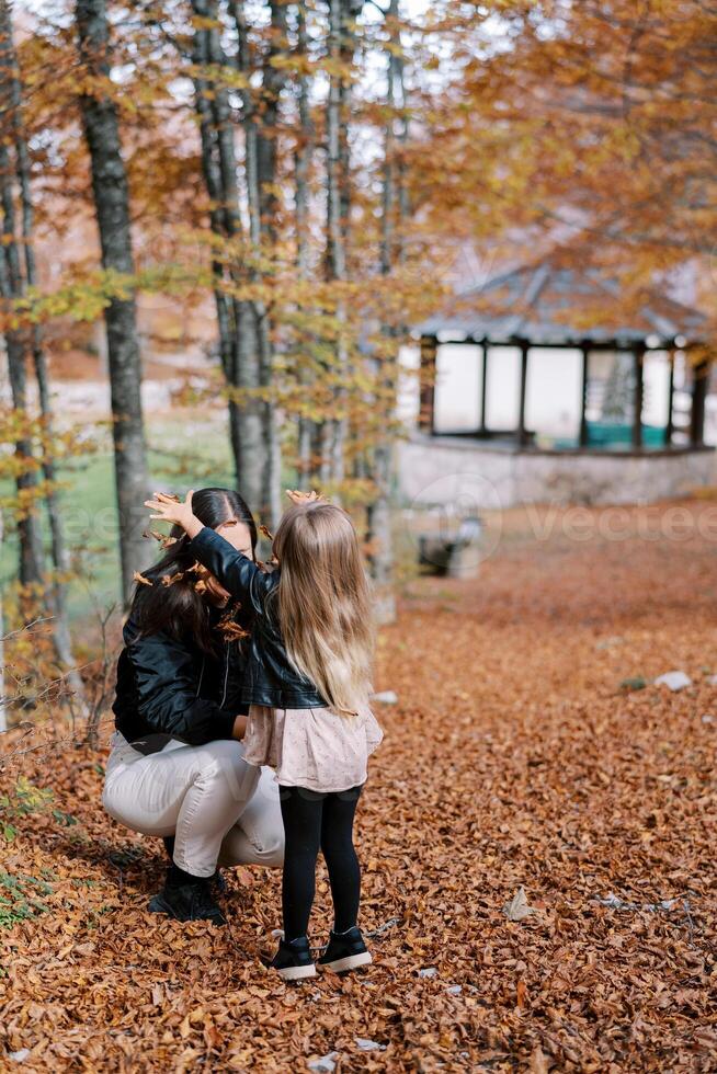 peu fille jette sec feuilles plus de sa mère squat dans un l'automne parc. retour vue photo