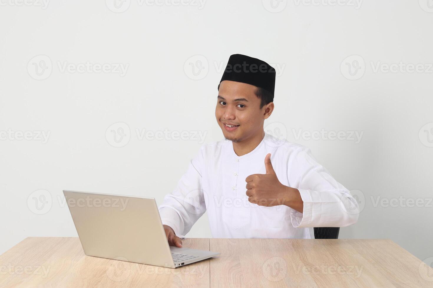 portrait de excité asiatique musulman homme dans koko chemise avec calotte travail sur le sien portable pendant jeûne sur Ramadan mois, montrant pouce en haut main geste. isolé image sur blanc Contexte photo