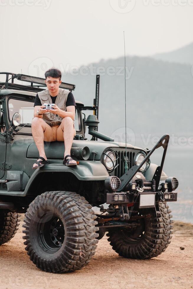 Jeune homme est assis sur une fortement modifié voiture, en portant une caméra, prêt pour un hors route la photographie aventure. photo