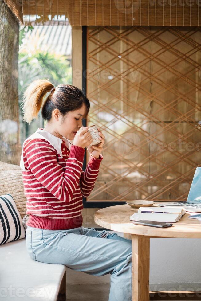 décontractée Jeune femme prise plaisir dans sirotant une chaud boisson tandis que en train de lire une livre dans une confortable, ensoleillé café paramètre. photo