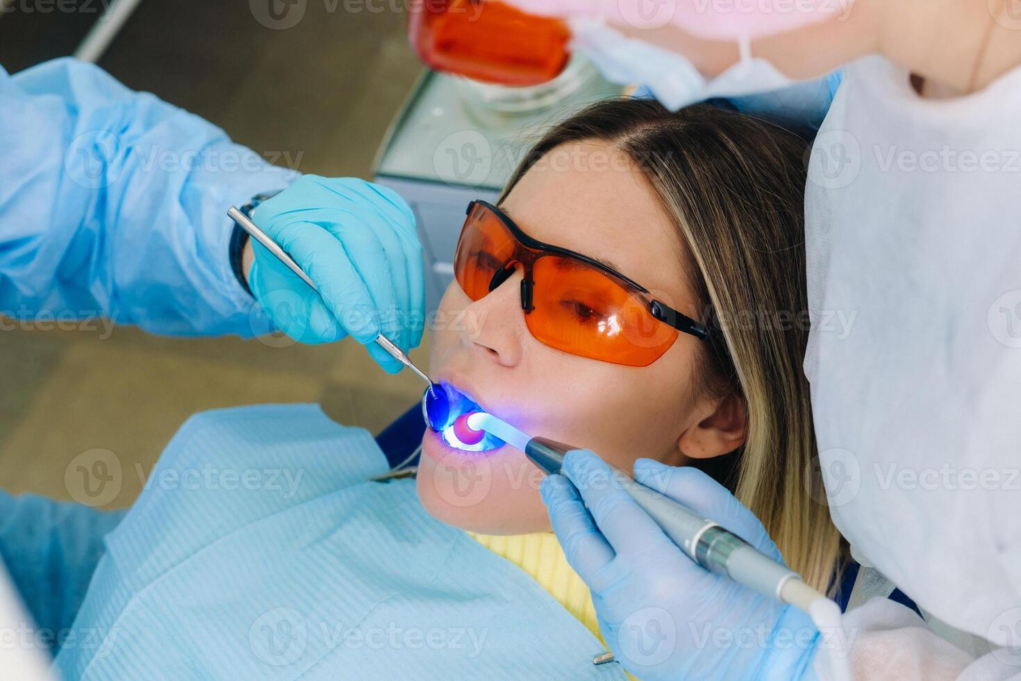 une Jeune magnifique fille dans dentaire des lunettes traite sa les dents à le dentiste avec ultra-violet lumière. remplissage de les dents photo