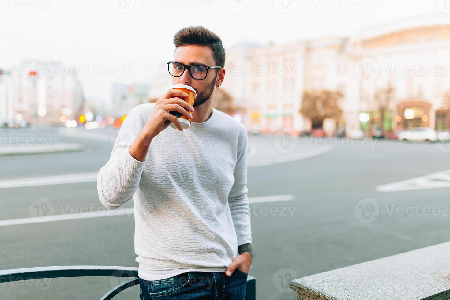 branché homme permanent avec à emporter café, souriant agréablement, en marchant sur e ville rue. content insouciant Beau gars dans lunettes photo