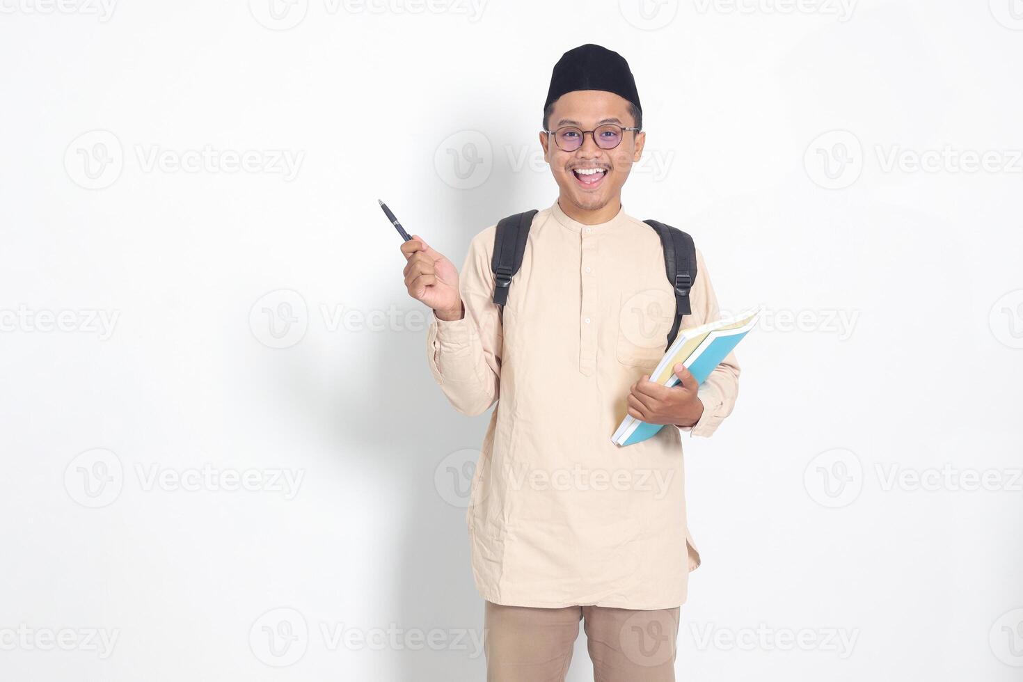 portrait de excité étudiant asiatique musulman homme dans koko chemise avec calotte porter sac à dos, en portant école livres, montrer du doigt à le côté. islamique éducation concept. isolé image sur blanc Contexte photo