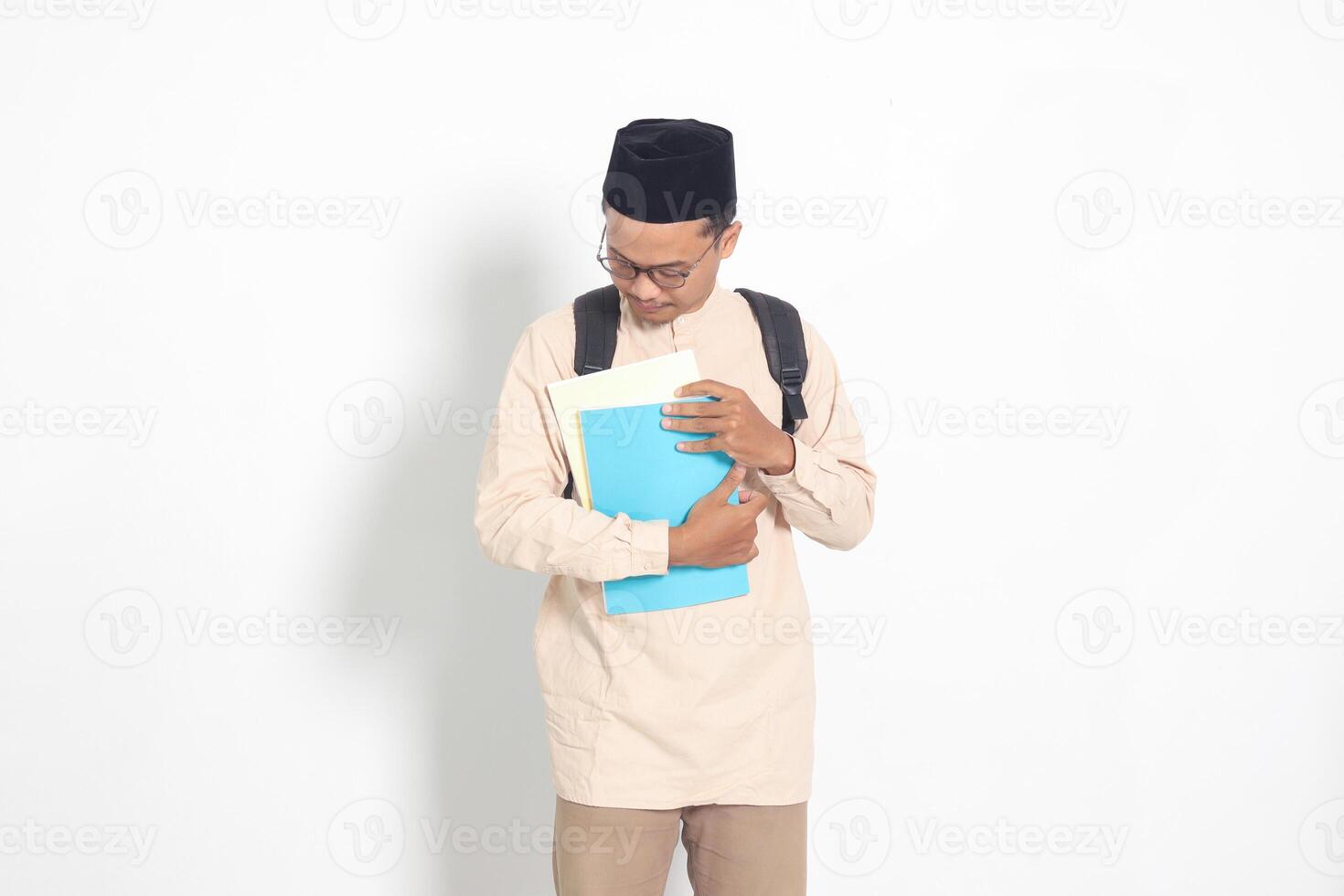 portrait de excité étudiant asiatique musulman homme dans koko chemise avec calotte porter sac à dos, tandis que en portant le sien école livres. islamique éducation concept. isolé image sur blanc Contexte photo