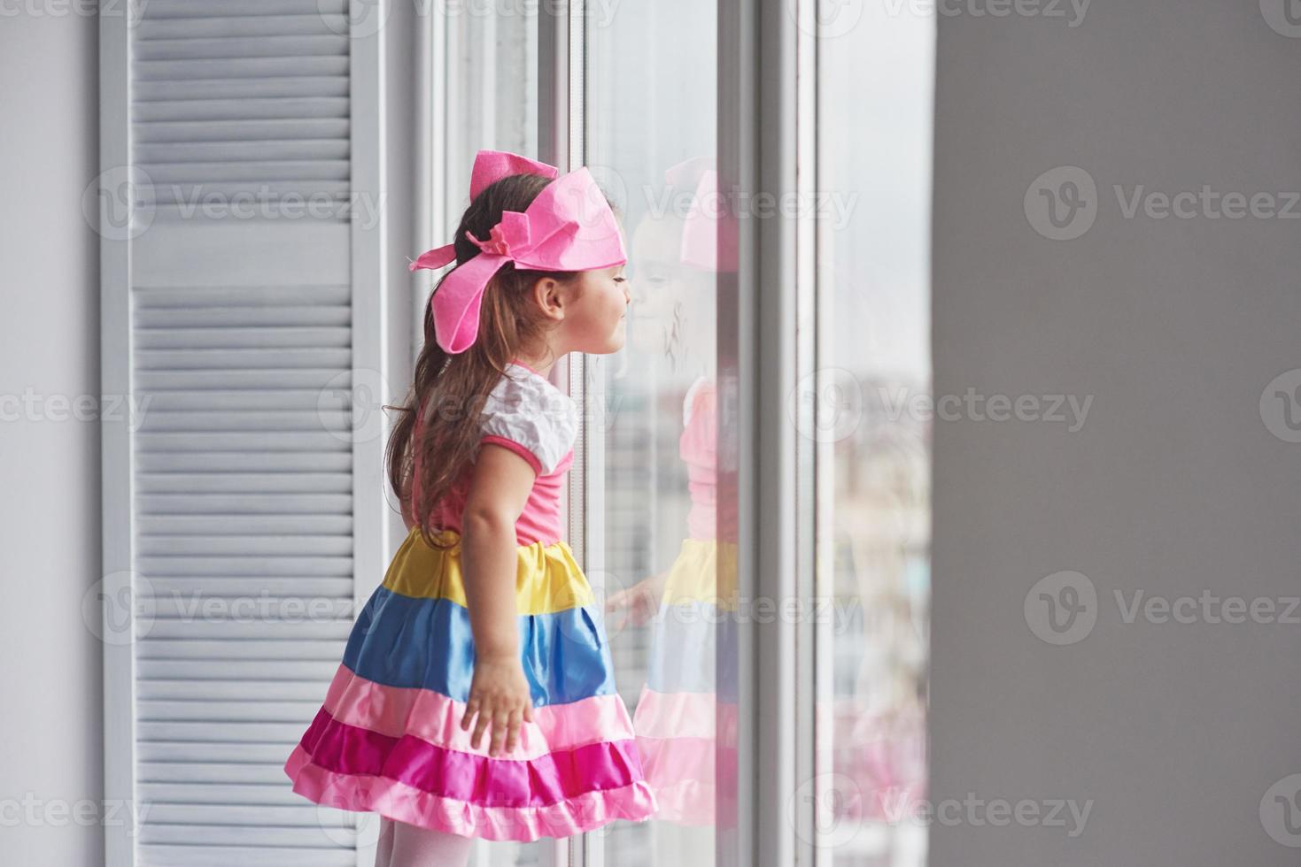 curiosité enfantine. photo de jeune fille mignonne en vêtements colorés debout près de la fenêtre et regardant à l'extérieur