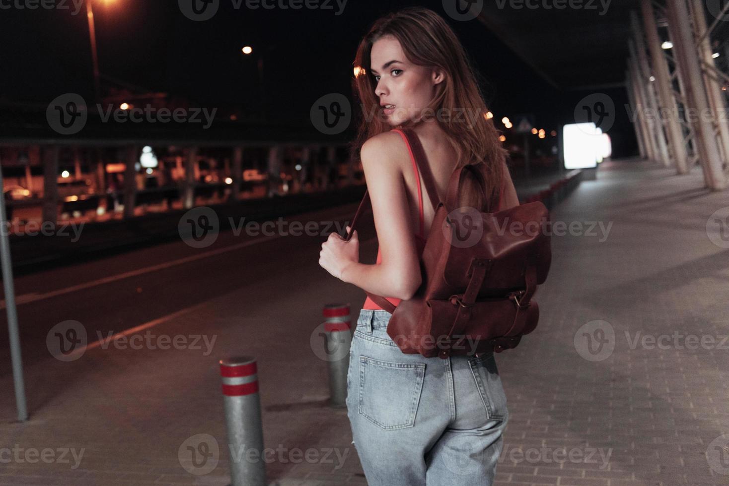 jolie jeune fille se tient sur le trottoir avec un sac sur le dos et regarde vers le bord de la route photo