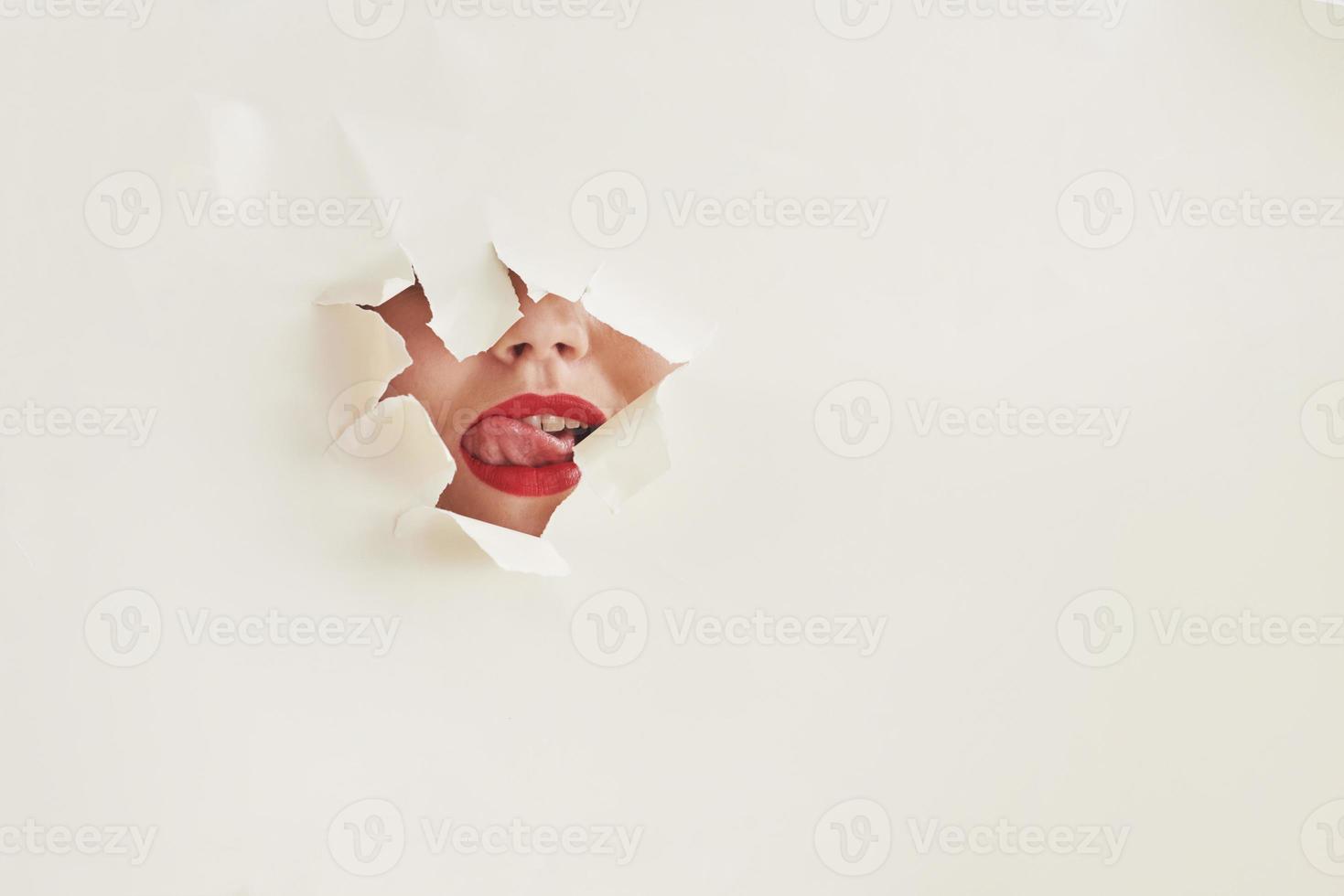 se lèche les lèvres. trou déchiré dans du papier blanc avec bouche de jeune femme photo