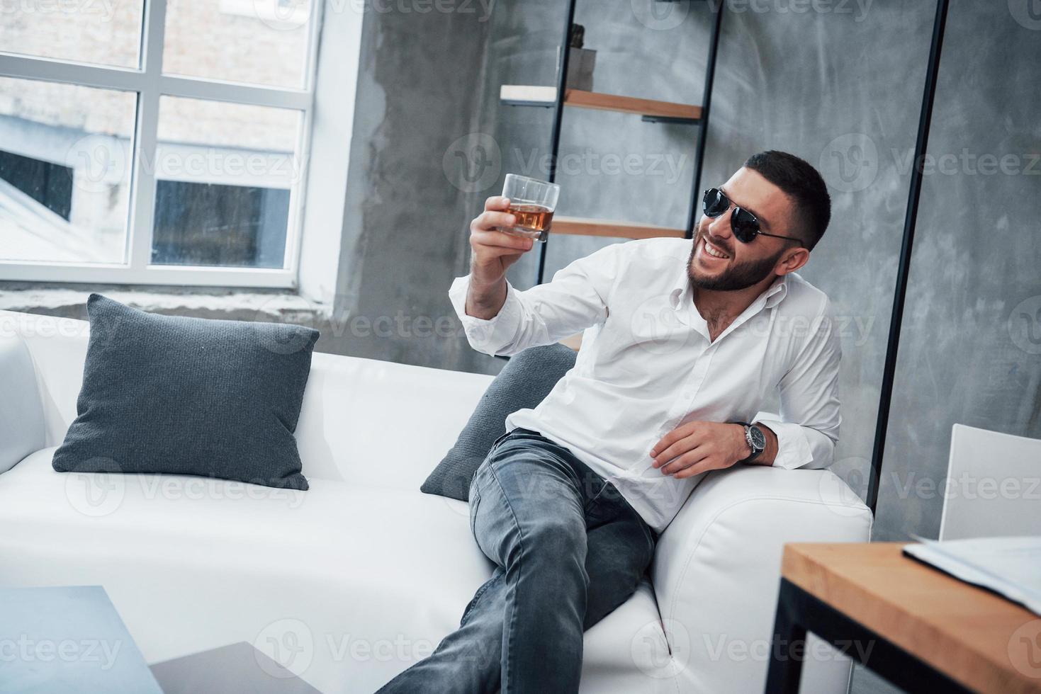 élever du whisky et sourire. jeune homme aux cheveux courts à lunettes de soleil assis sur un canapé au bureau photo