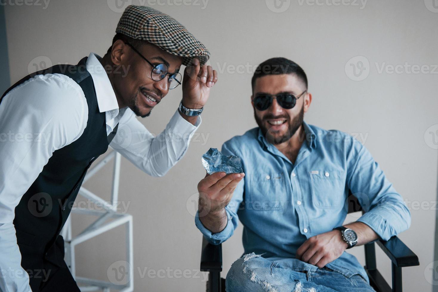 un mec aux lunettes de soleil tient la pierre à bijoux lorsqu'un homme afro-américain touche une casquette. photo d'amis métis souriants ont une séance photo en studio