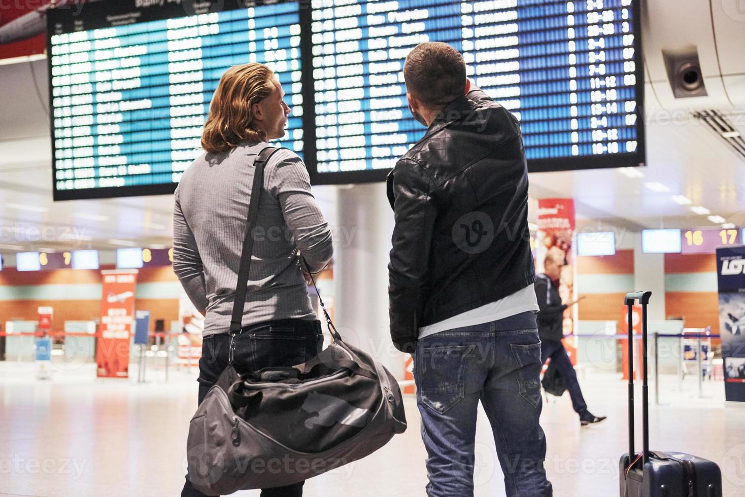 l'homme montre l'heure d'arrivée de l'avion. photo de deux camarades se trouvant à l'aéroport près du système d'affichage des informations de vol