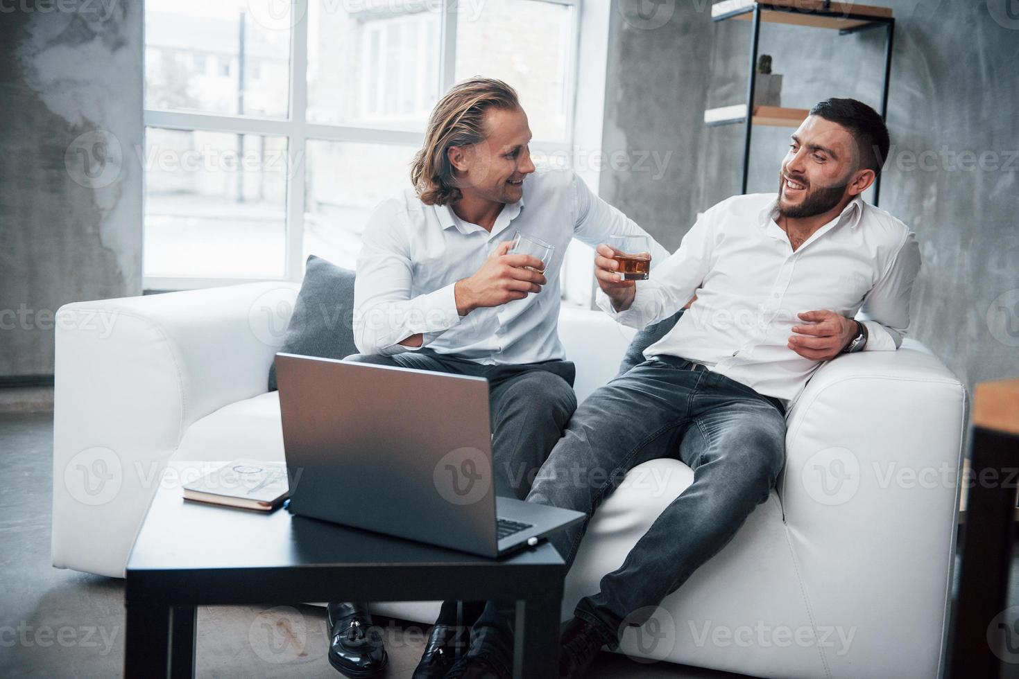 célébrer le succès. deux hommes d'affaires assis devant un ordinateur portable parlant de leurs plans et buvant du whisky photo