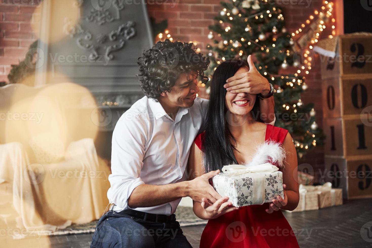 homme faisant une surprise pour sa petite amie. donne le nouvel an cadeau. jeune couple est assis sur le sol de la chambre de la chambre décorée de vacances photo