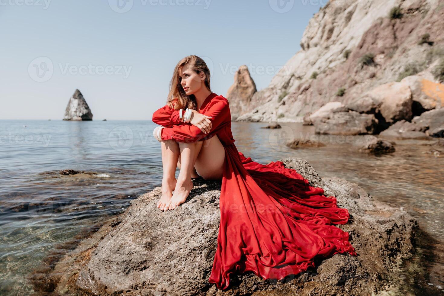 femme Voyage mer. content touristique dans rouge robe prendre plaisir prise image en plein air pour souvenirs. femme voyageur posant sur le Roche à mer baie entouré par volcanique montagnes, partage Voyage aventure périple photo