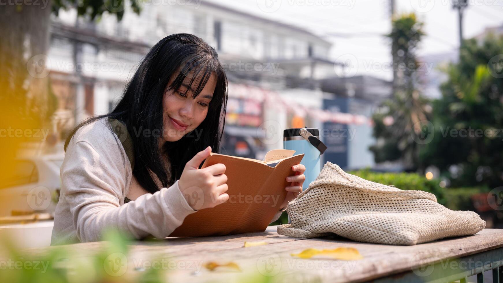 un attractif, content asiatique femme est en train de lire une livre à une table en plein air dans le ville. photo