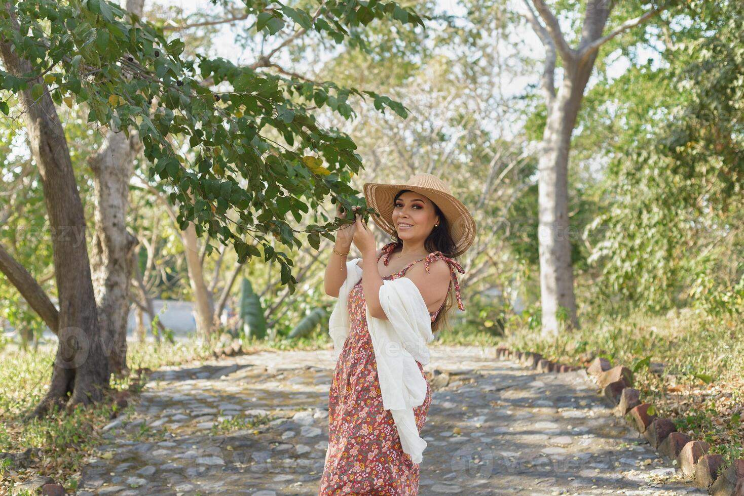 femme portant chapeau et robe avec fleurs admiratif la nature dans une Publique parc. ensoleillé été journée. photo