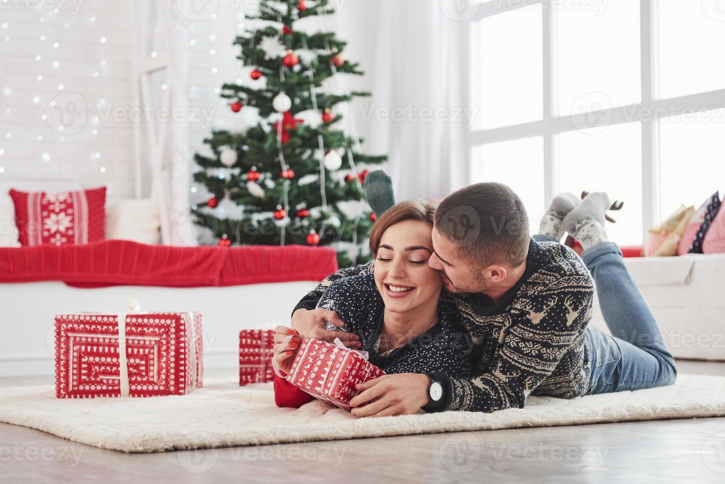 guy donne à sa femme un cadeau de Noël. beau jeune couple allongé sur le salon avec arbre de vacances vert à l'arrière-plan photo
