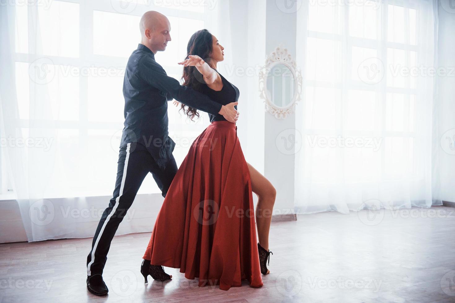 grande performance professionnelle. Jeune jolie femme en vêtements rouges et noirs dansant avec un homme chauve dans la salle blanche photo