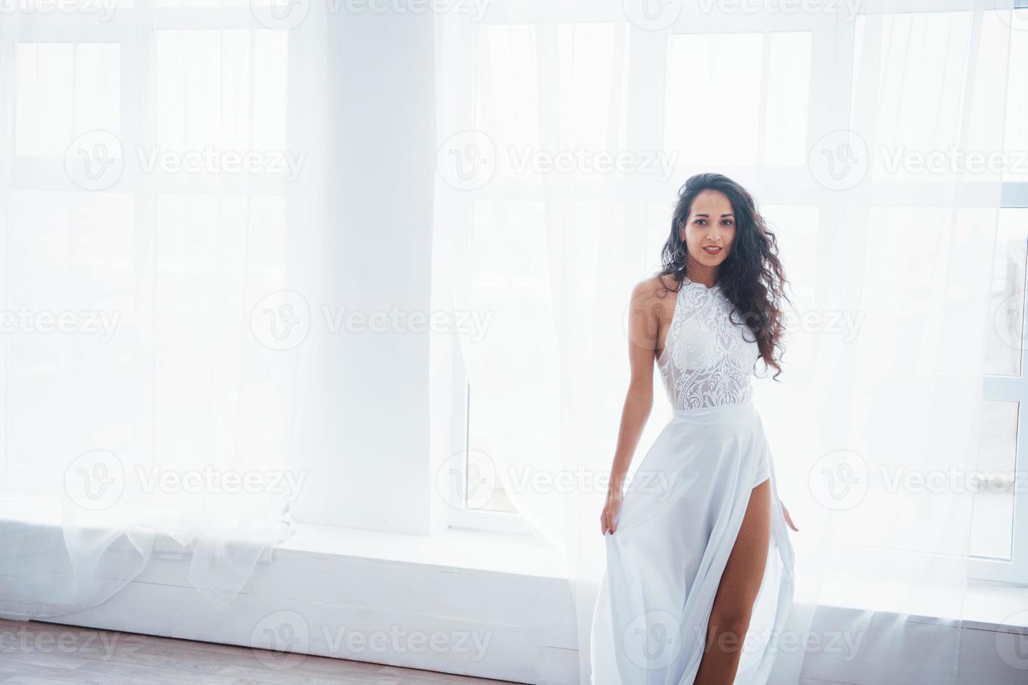 regardant droit dans la caméra. belle femme en robe blanche se tient dans une pièce blanche avec la lumière du jour à travers les fenêtres photo