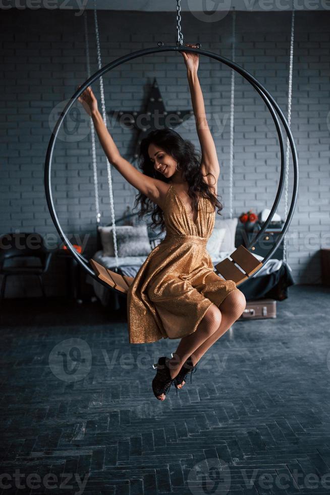 les mains sont levées. jolie femme en robe dorée est assise sur l'élégant banc en forme de cercle sur la chaîne. chambre de luxe photo