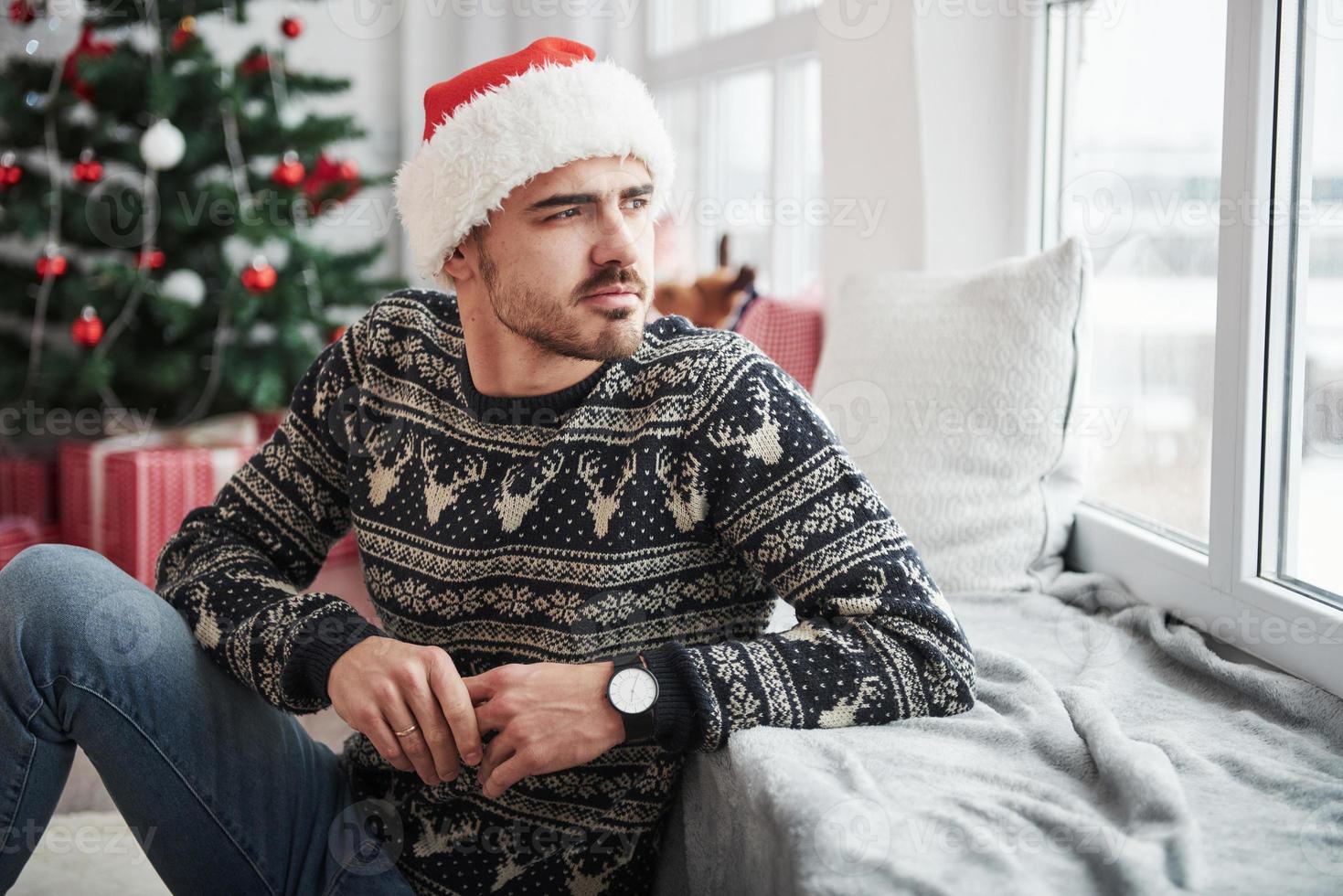 appuyé sur le rebord de la fenêtre. photo d'un homme en bonnet de noel et vêtements de vacances regarde par la fenêtre. arbre de noël sur fond