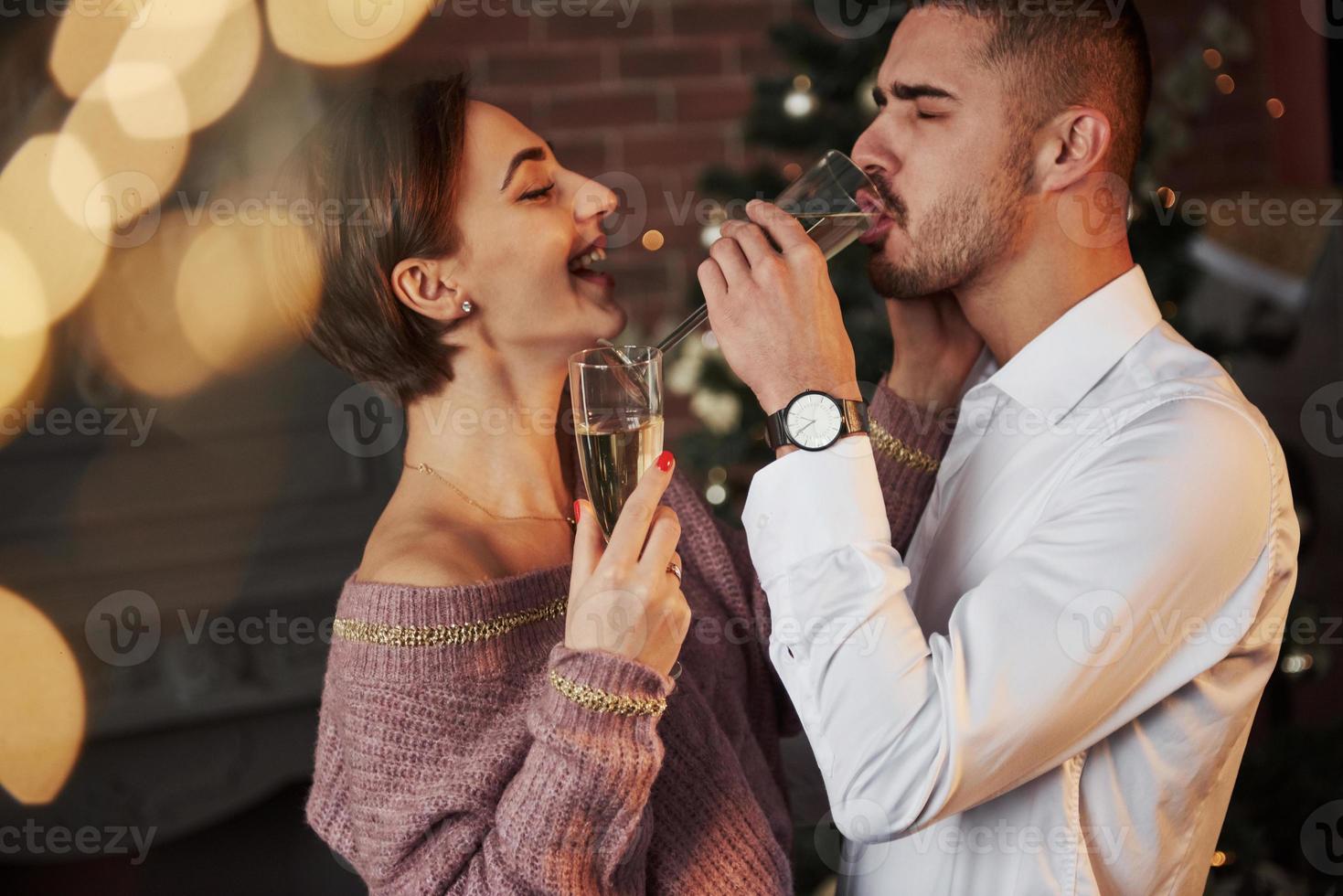 mec boit du champagne pendant que sa petite amie rit. joli couple célébrant le nouvel an à l'intérieur avec de beaux vêtements classiques sur eux photo
