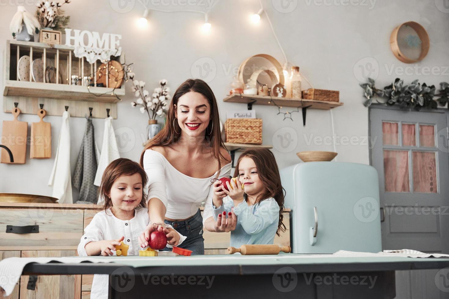 belle jeune femme nourrit deux enfants avec des pommes alors qu'ils sont assis près de la table avec des jouets photo