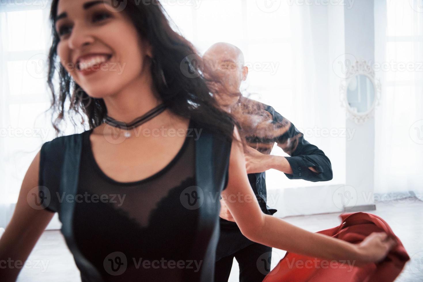 Couple heureux. Jeune jolie femme en vêtements rouges et noirs dansant avec un homme chauve dans la salle blanche photo
